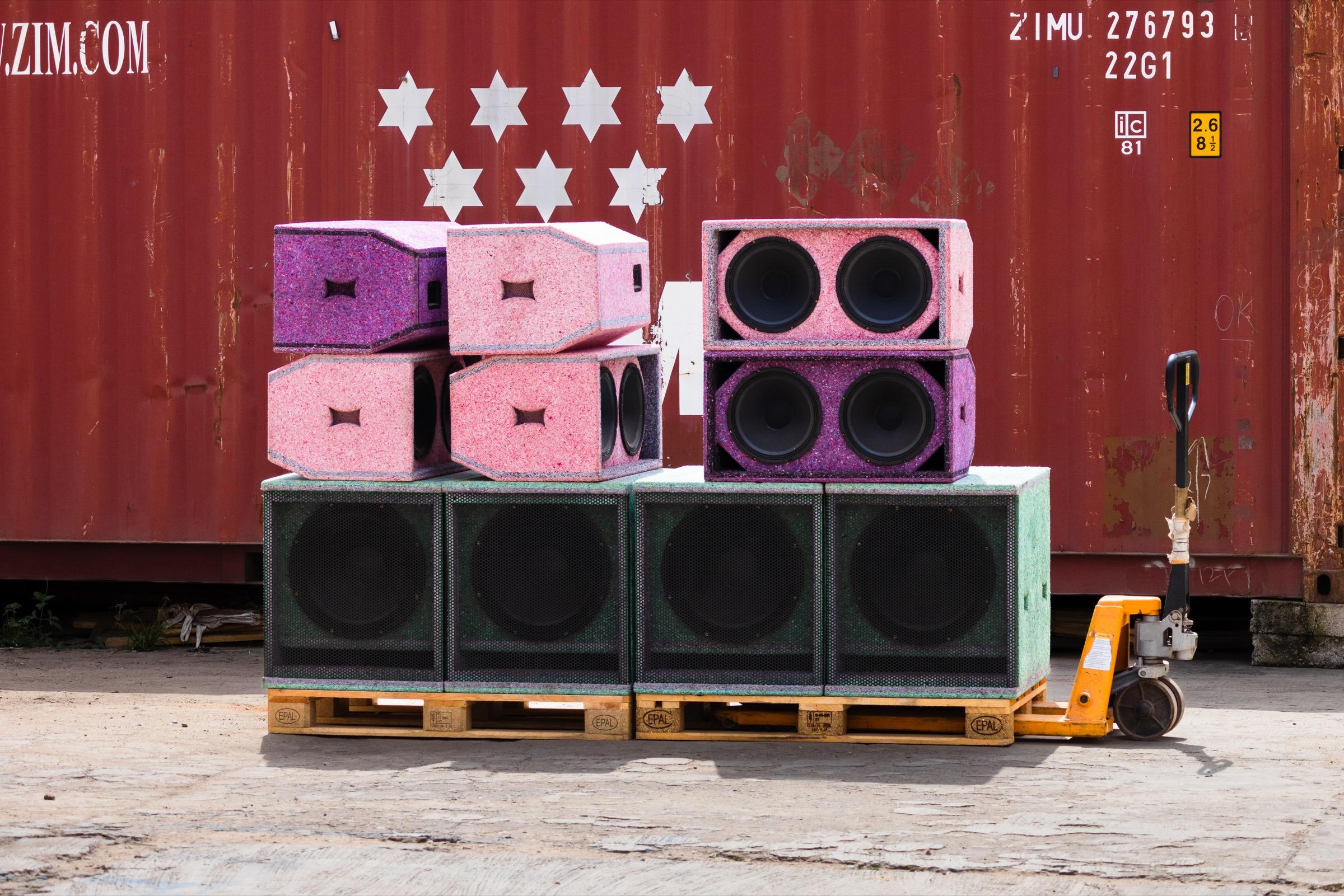 Speaker cabinets moulded from pink, purple and green recycled plastic, stacked on pallets in front of a red shipping container.